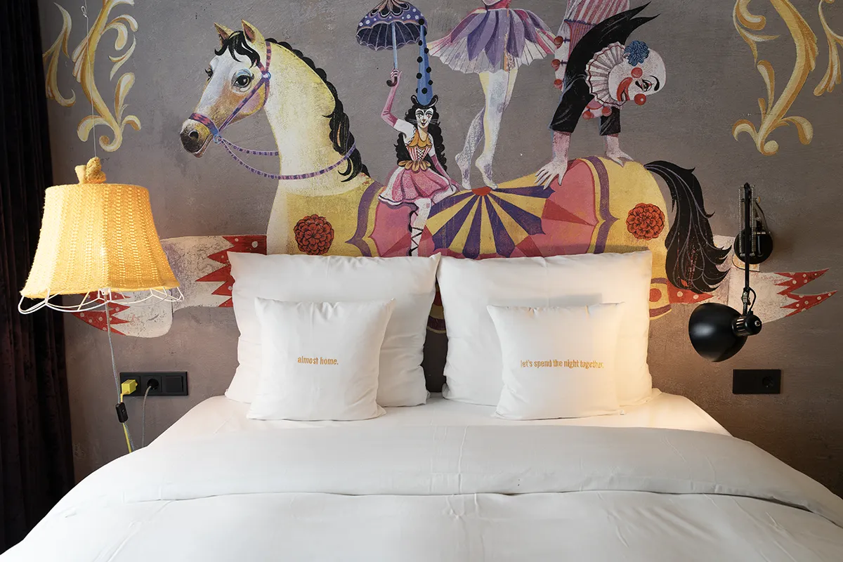A freshly made hotel bed with a circus-themed painting on the wall behind it.